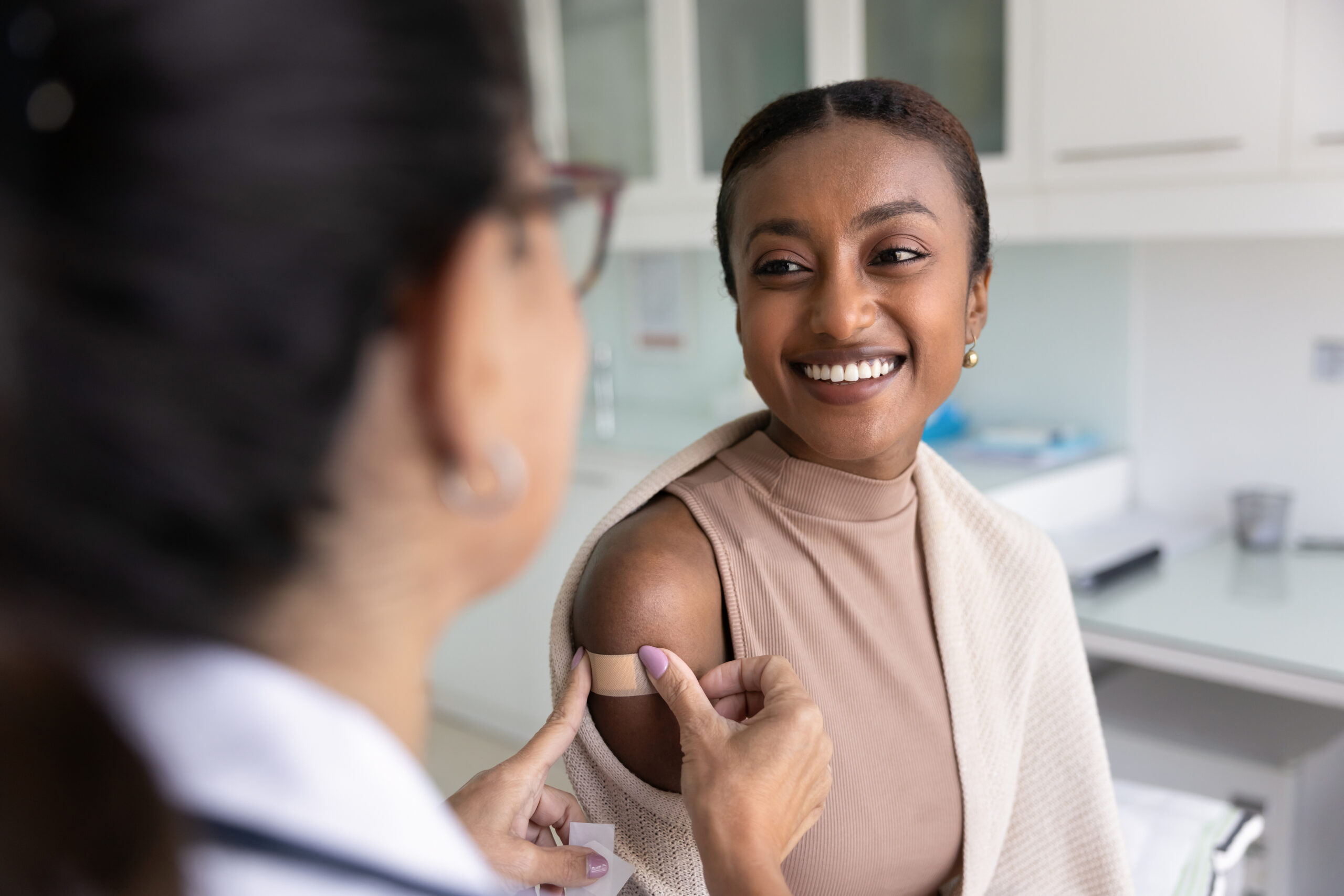 Cheerful young patient woman visiting doctor for vaccination, talking to physician, nurse sticking plaster on shoulder after injection, smiling, getting vaccine for protection from flu virus